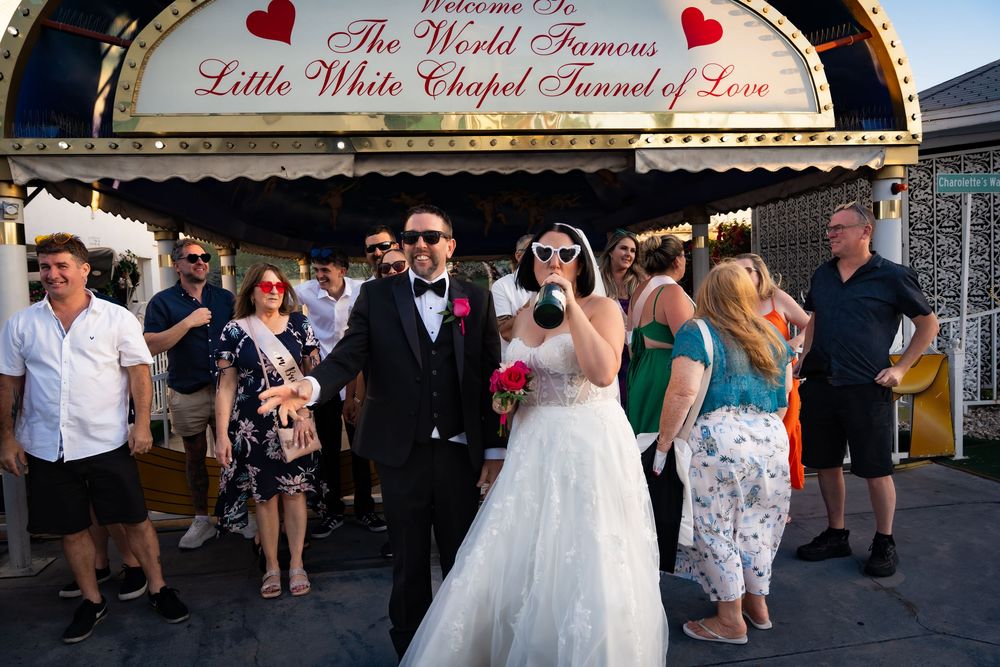 A couple poses outside the famous Little White Chapel wedding venue in Las Vegas with guests standing nearby.