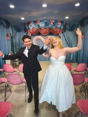 Happy newlyweds dancing and celebrating in a reception hall decorated with blue curtains and pink chairs.