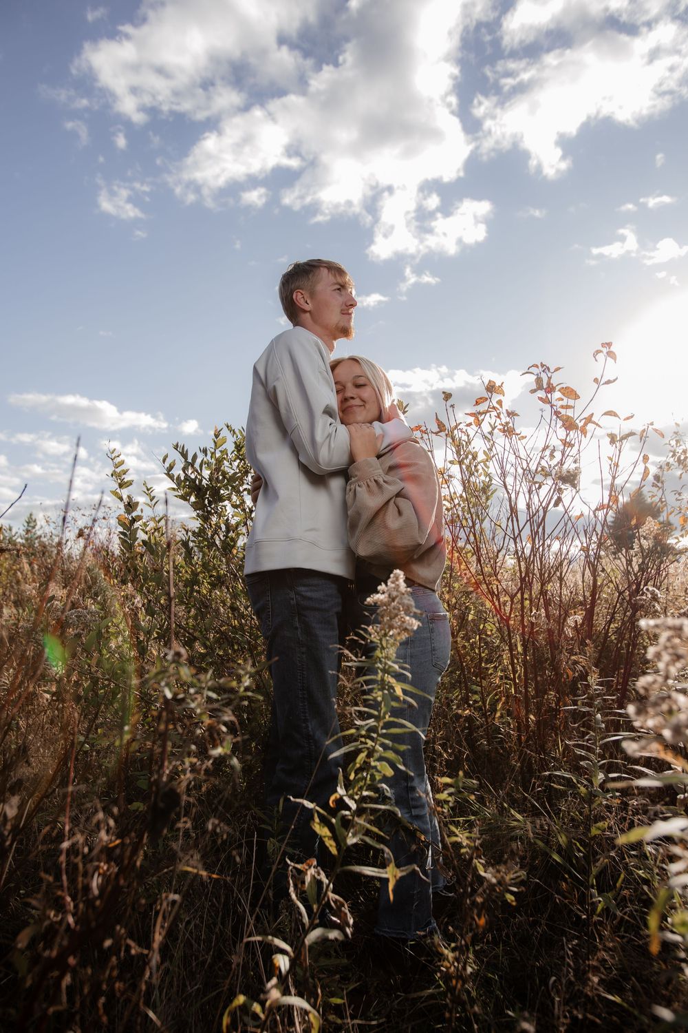 Couple embracing in tall grass field against bright cloudy sky.
