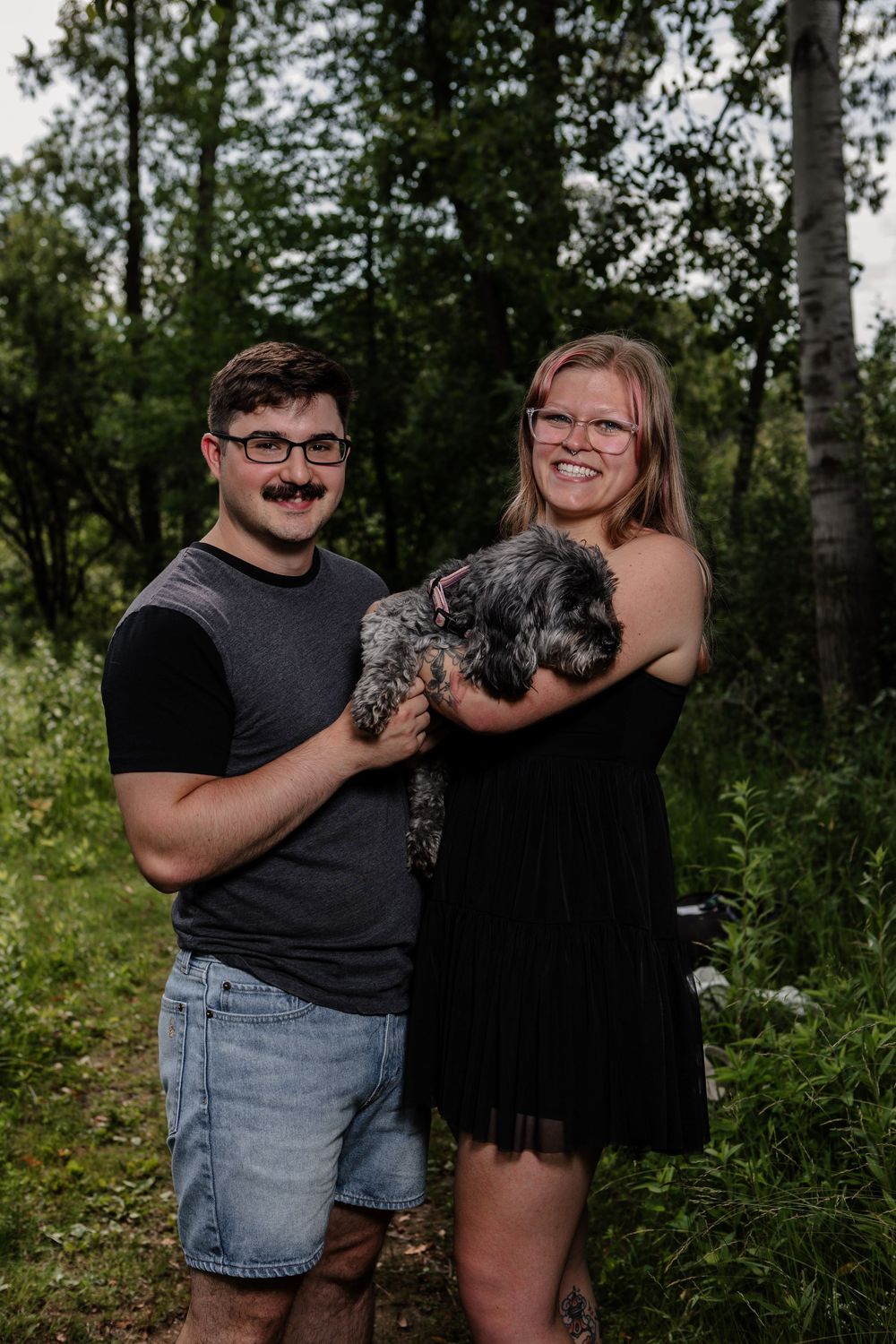 A couple holds their small dog while posing in a wooded outdoor setting.