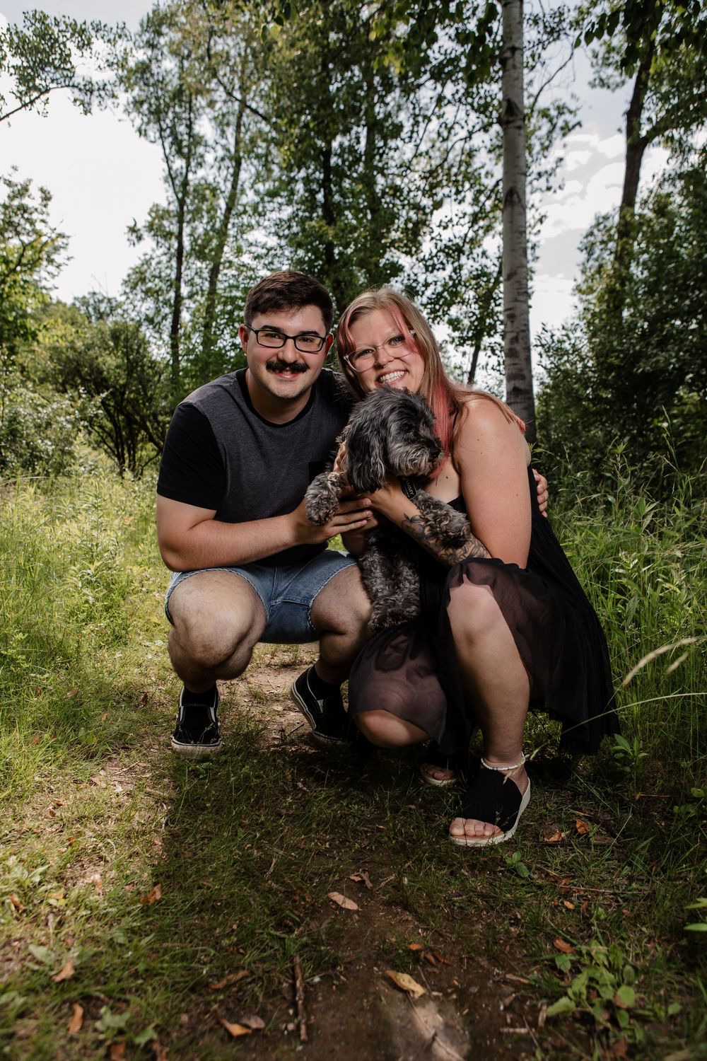 A couple crouches with their pet dog for photos in a natural forest setting.