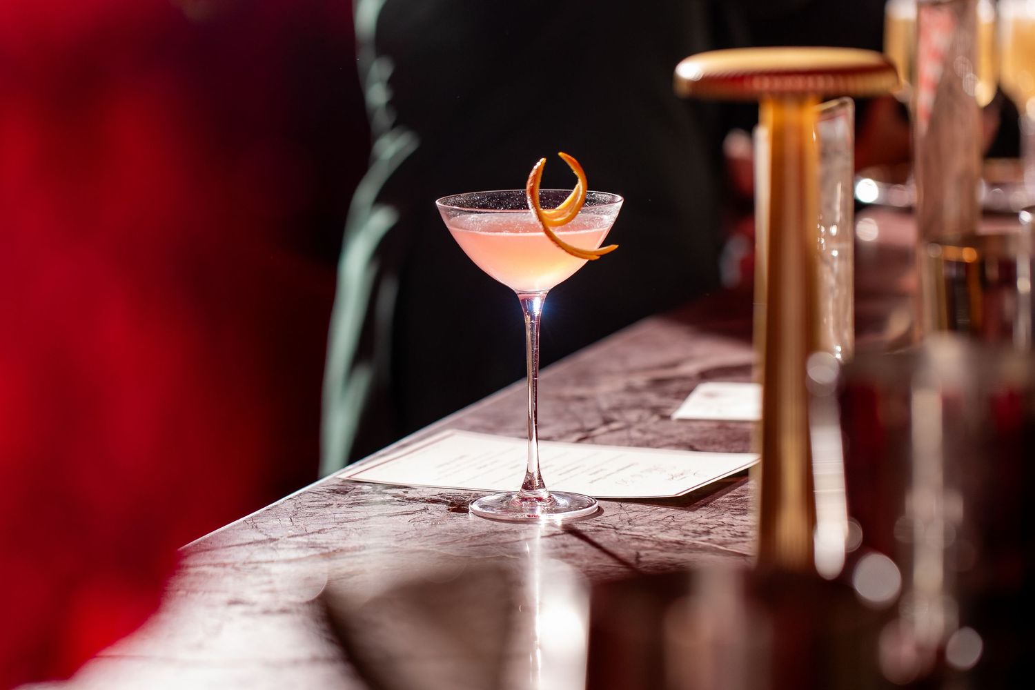A pink cocktail in a champagne glass sits on a bar with orange rind on the rim - Event Photography