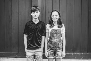 Two young people stand against a dark wooden wall, one wearing overalls and one in a black polo shirt, in black and white.