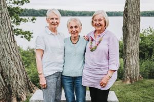 Three seniors stand together smiling outdoors with a lake and trees visible in the background on a sunny day.