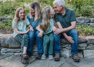 A family sits together and laughs while relaxing on a stone wall surrounded by greenery on a sunny day.