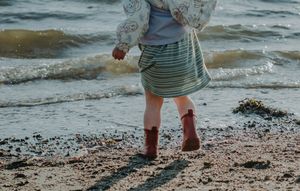 A young girl in a striped skirt and red boots is playing on a sandy beach near ocean waves at Mitchell Field in Harpswell, Maine.