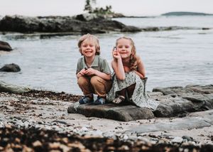 Two happy children smiling and posing together on a rocky beach in Boothbay, Maine, with ocean and islands in the background at Ocean Point.
