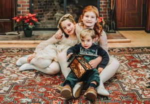 Three young siblings pose together on a red rug by a fireplace in holiday attire.  They became a family through foster care and adoption.