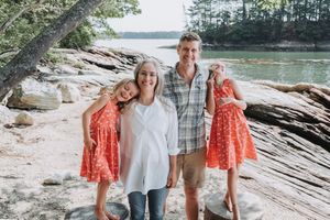 A family wearing casual clothes poses together on a rocky beach beside water and trees at Wolfe's Neck Park in Freeport, Maine.