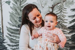 A mother holds her happy baby girl in a floral outfit near a painted forest mural located at the Wolfe's Neck Education Center in Freeport.