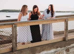 Three sisters lean against a wooden railing while enjoying a scenic waterfront view on a sunny day.