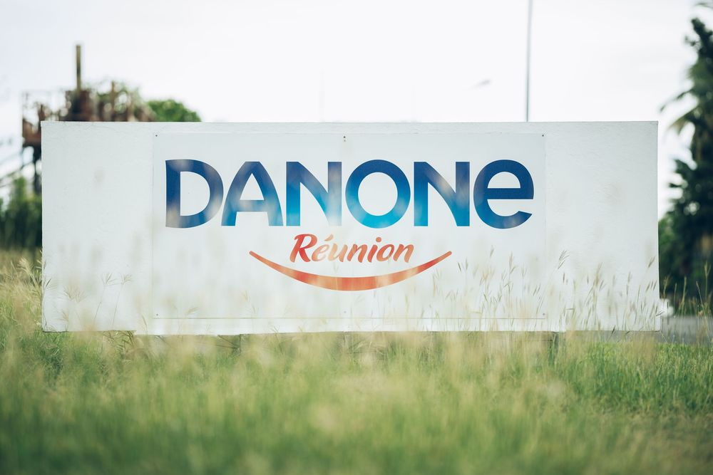 A Danone company logo sign mounted on a white wall in a grassy outdoor setting with blurred background.