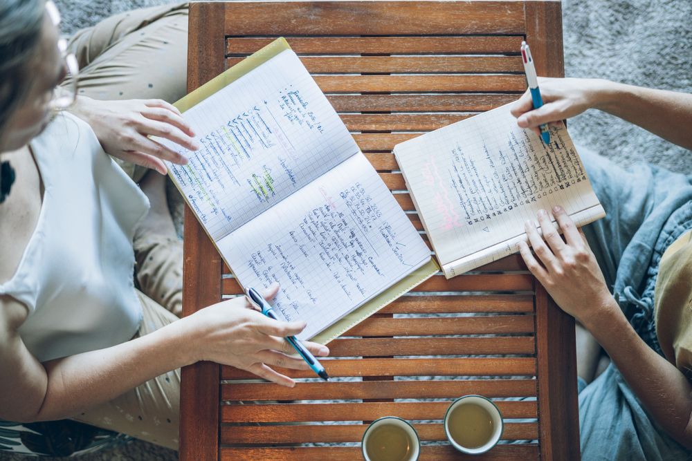 Two people study and discuss notes together at a wooden table with coffee cups.