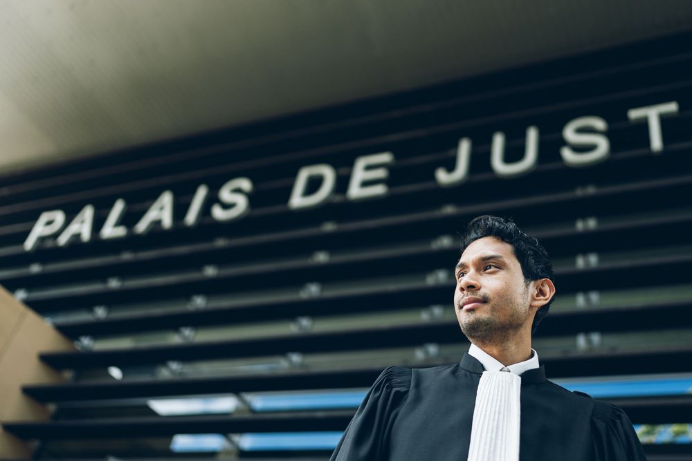 A lawyer in traditional court attire stands in front of the Palais de Justice building.