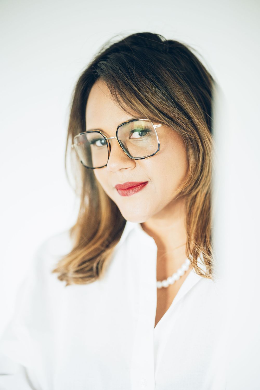 Portrait of someone with ombre brown hair wearing rectangular glasses and a white blouse with pearl necklace.