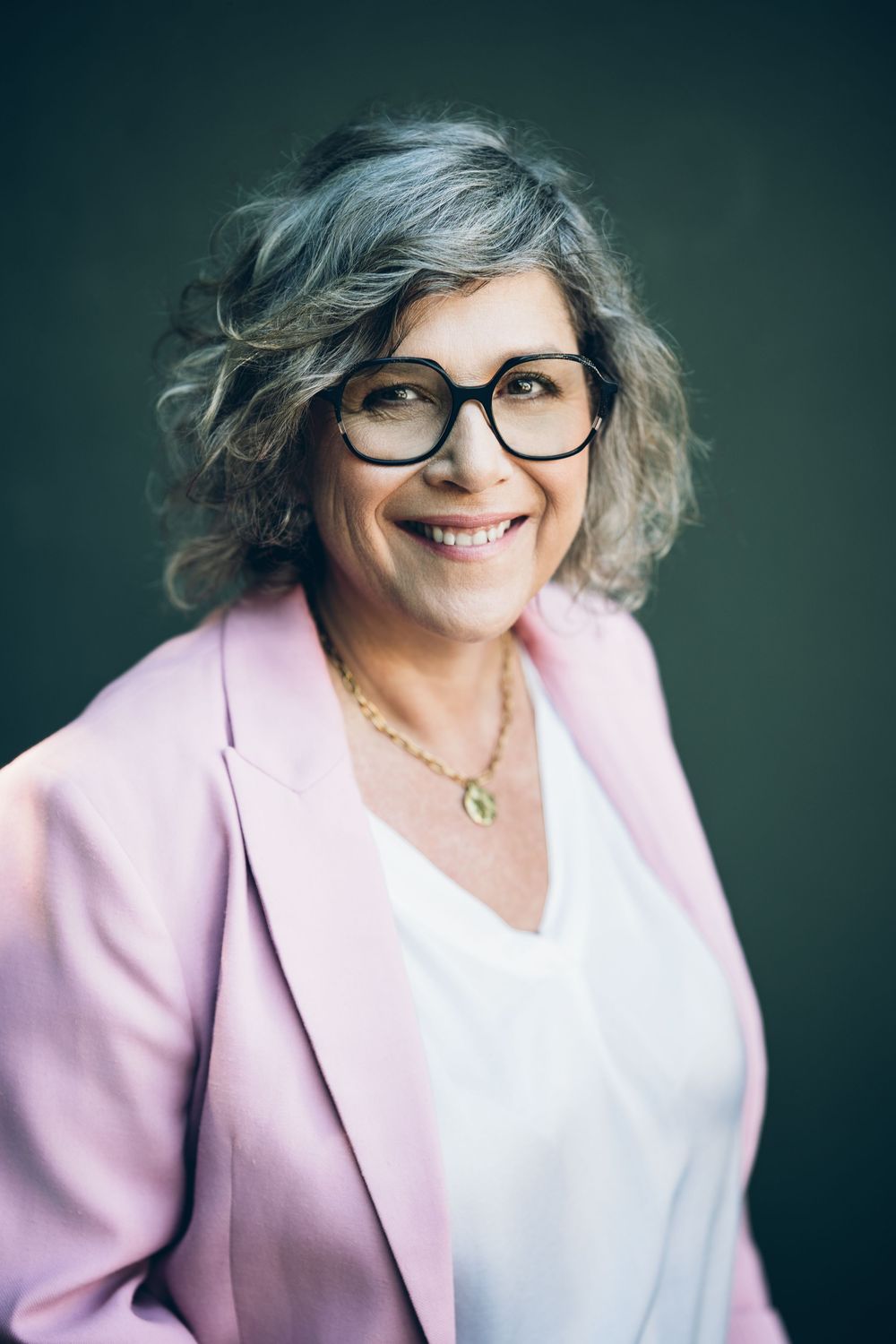 Professional studio portraits show a joyful senior woman in pink blazer and glasses against dark background.
