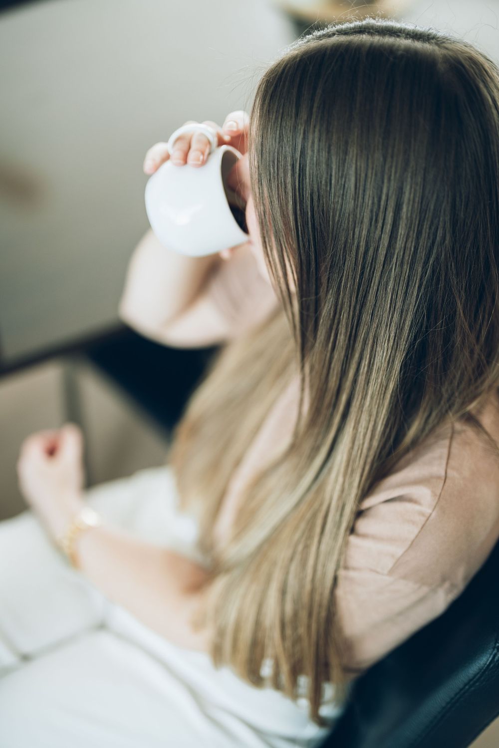 Woman with long straight ombre hair drinking from a white coffee cup while sitting in a chair.