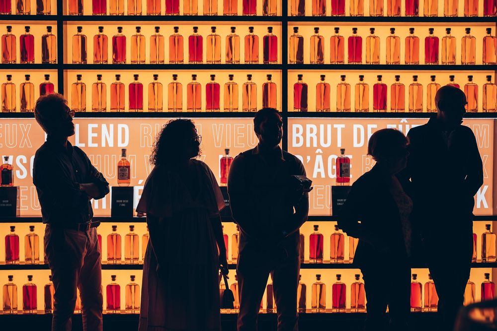 Silhouettes of people against glowing amber bar shelves filled with illuminated liquor bottles at a dimly lit establishment.
