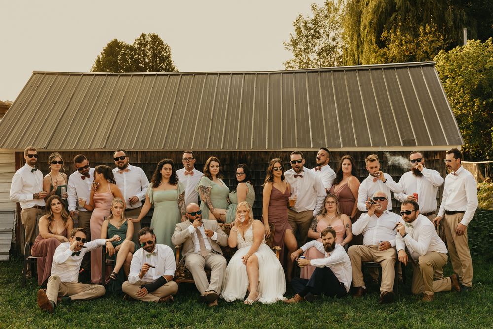 Large wedding party poses together on rustic barn grounds during sunset.