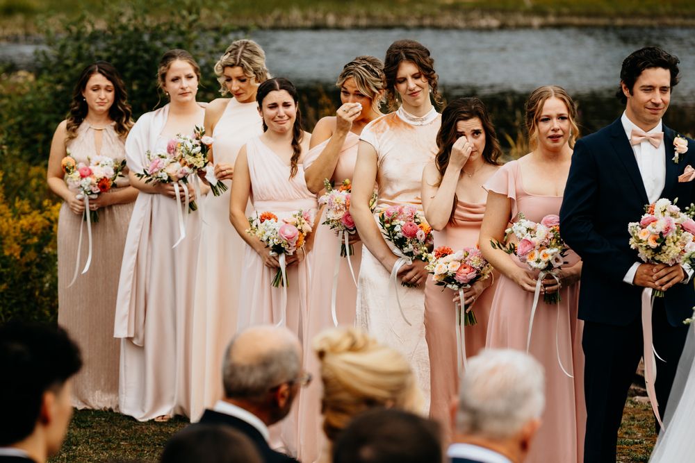 Wedding party members in blush pink dresses hold colorful bouquets while standing outdoors.