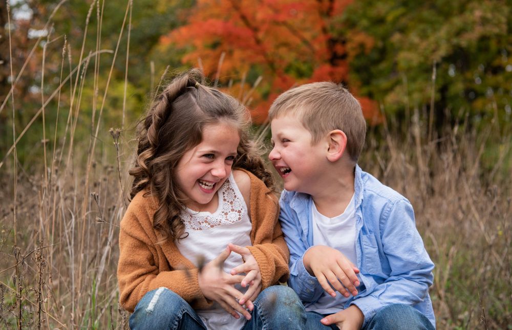 Two children laughing together outdoors during autumn with vibrant orange foliage in the background.