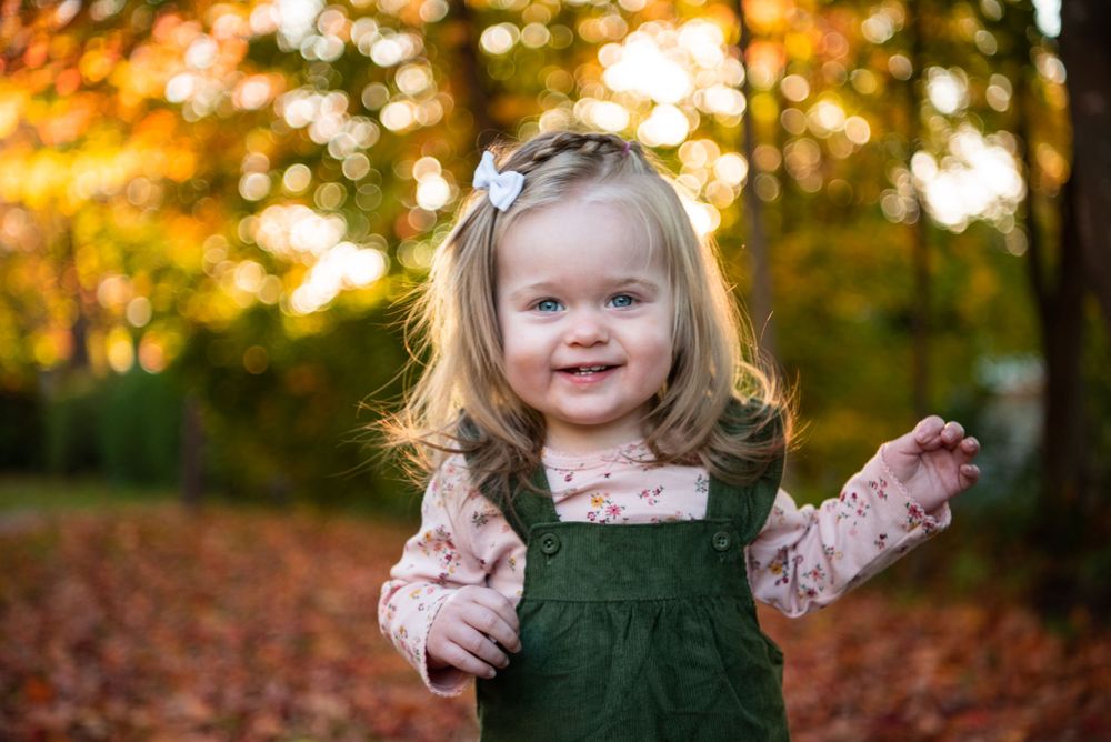 A child in a green corduroy dress and floral shirt stands among fallen autumn leaves with golden bokeh in background.