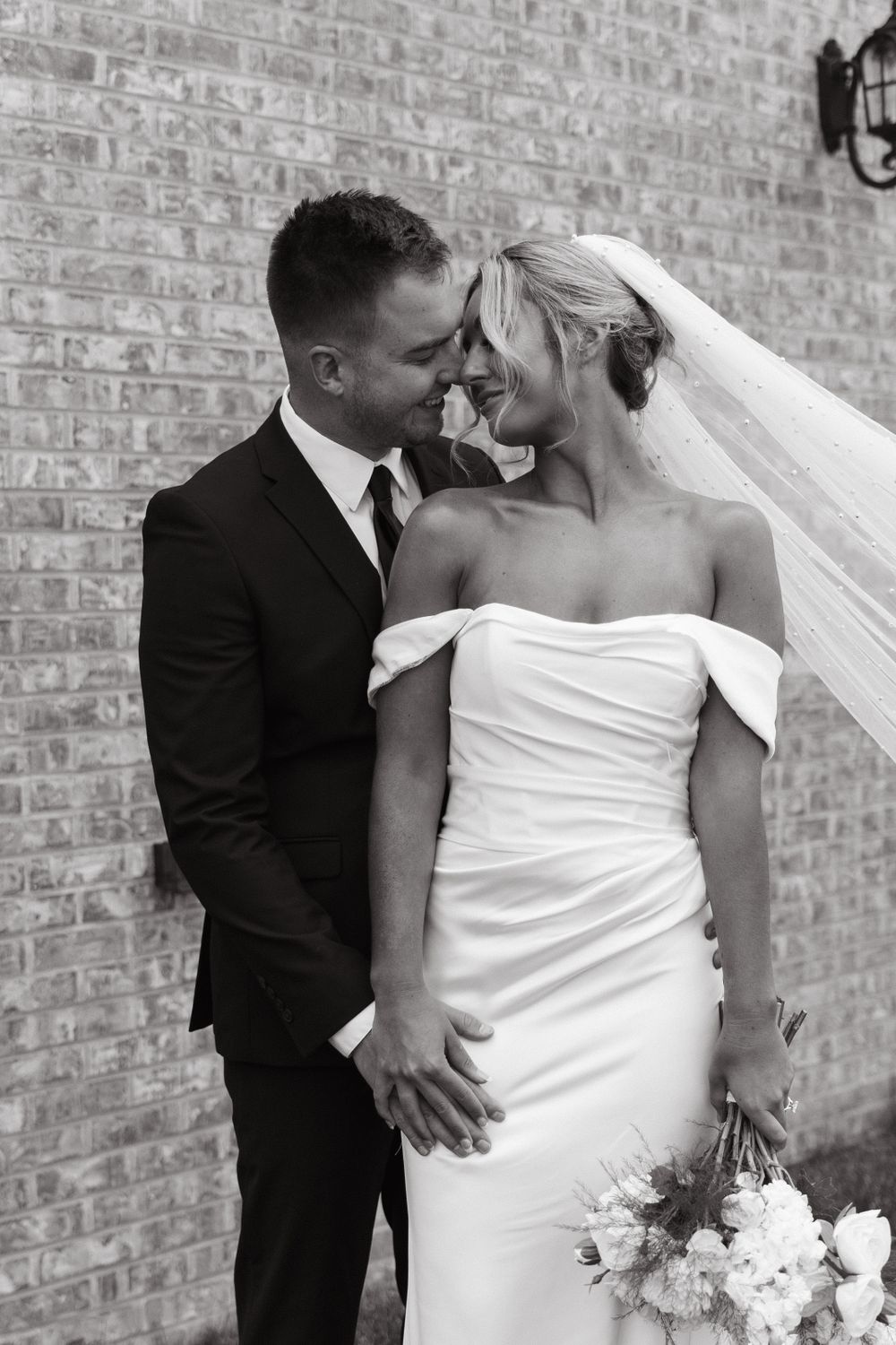 Romantic black and white photo of newlyweds sharing an intimate moment against a brick wall.