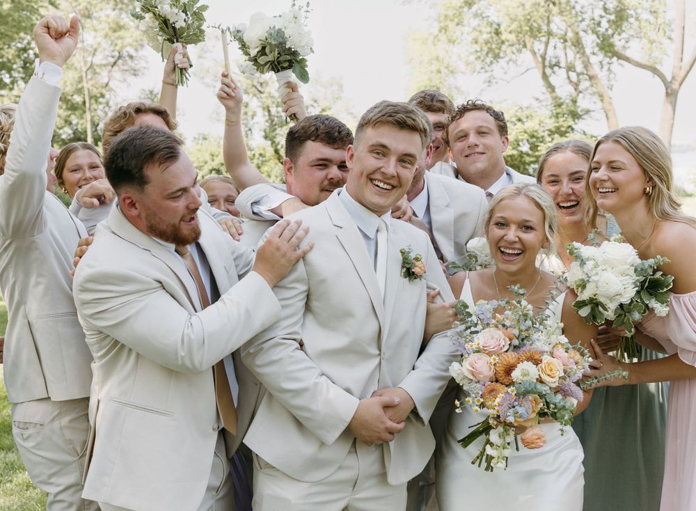 Wedding party celebrates joyfully together outdoors wearing light colored attire.