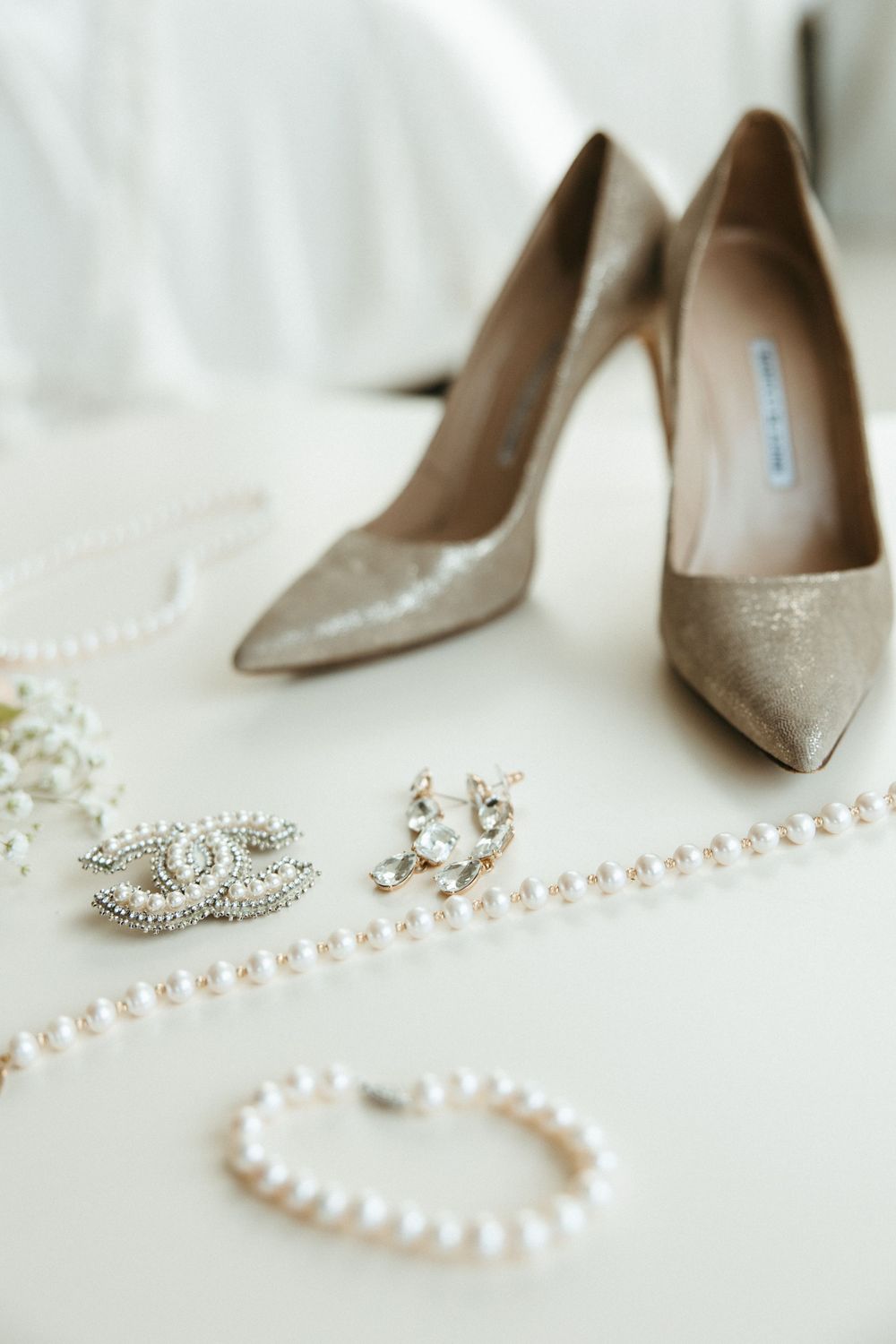 Wedding accessories including gold heels, pearl jewelry and crystal earrings arranged on white fabric.