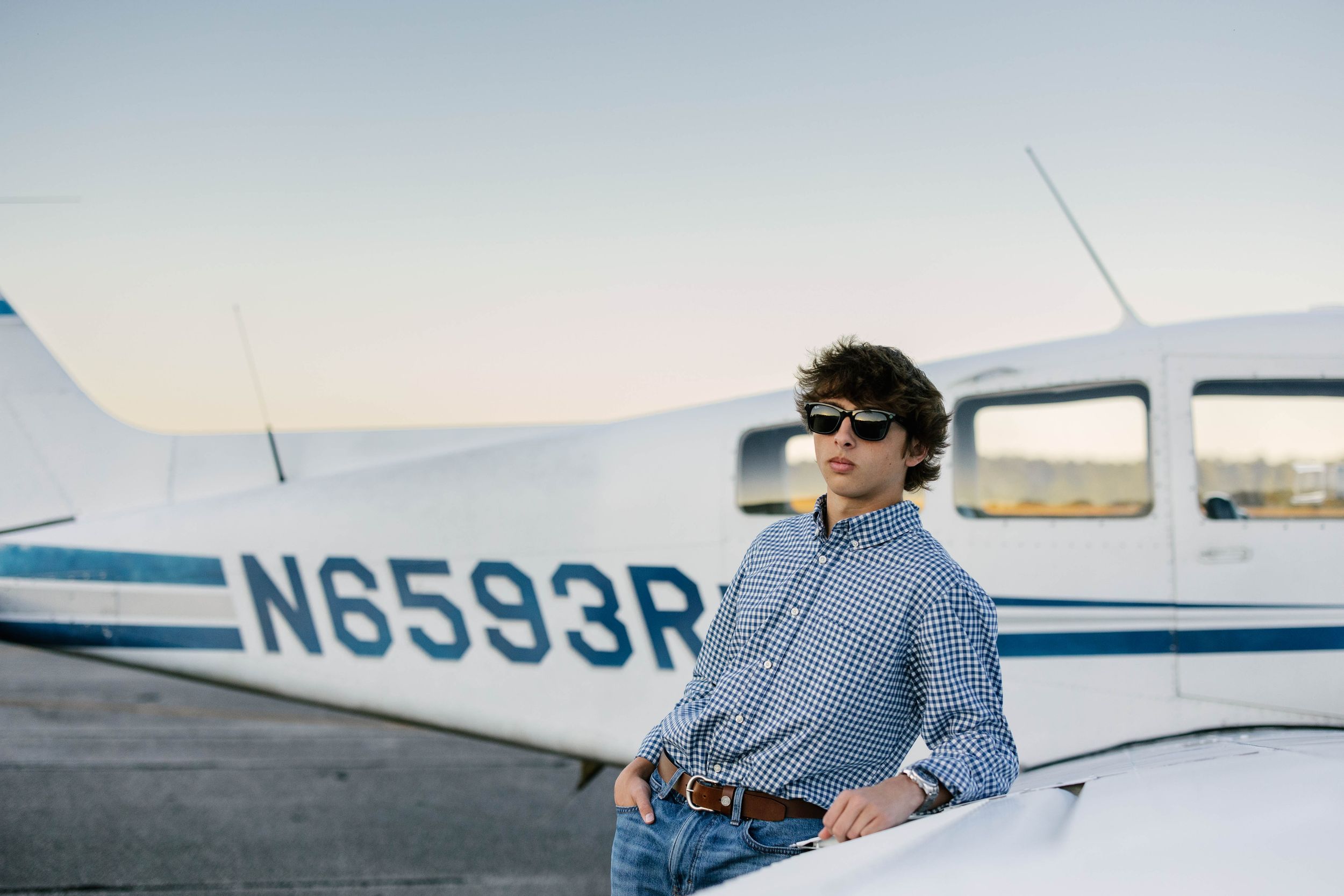 A person wearing sunglasses and a striped shirt stands next to a small private aircraft on a sunny day.
