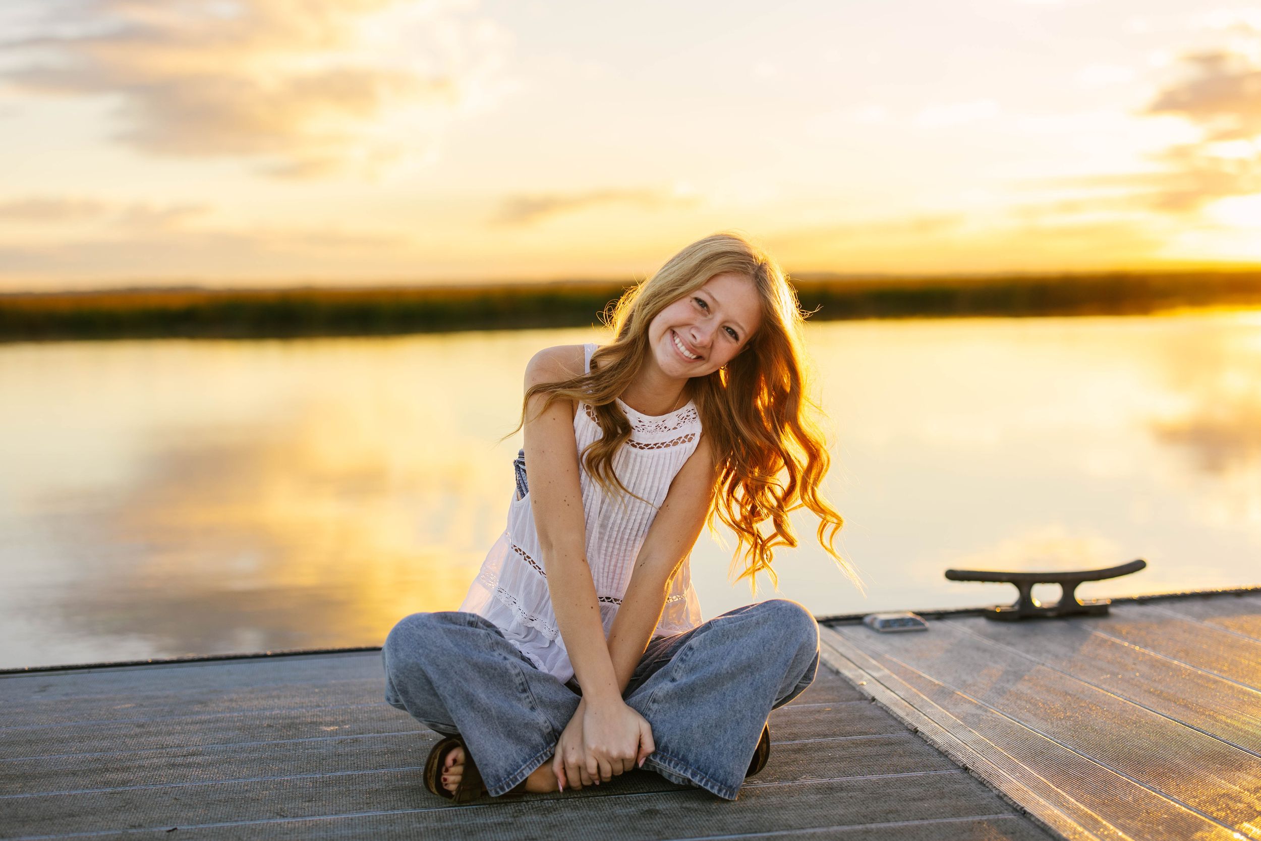 Person sitting cross-legged on wooden dock at sunset with golden light reflecting on calm lake waters.