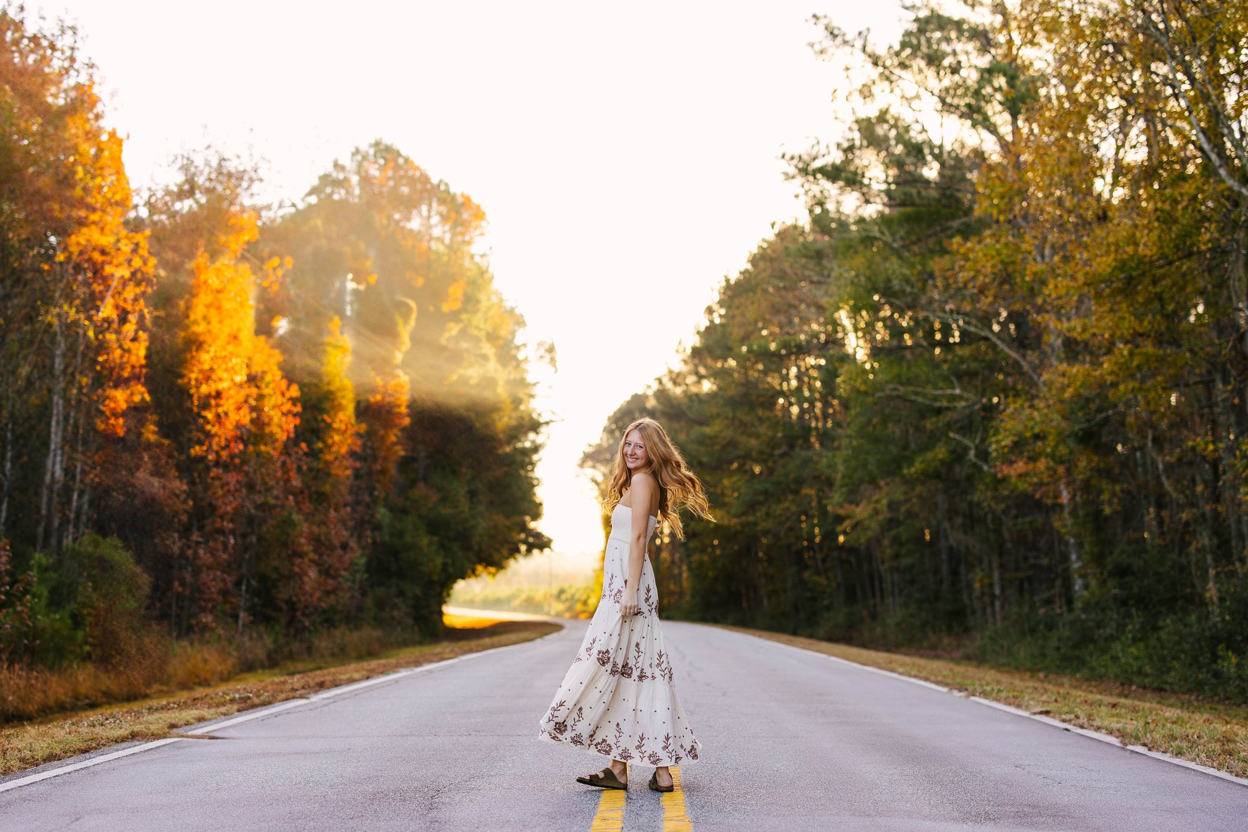 Person in flowing dress twirling on empty rural road with fall foliage and sunlight filtering through trees.