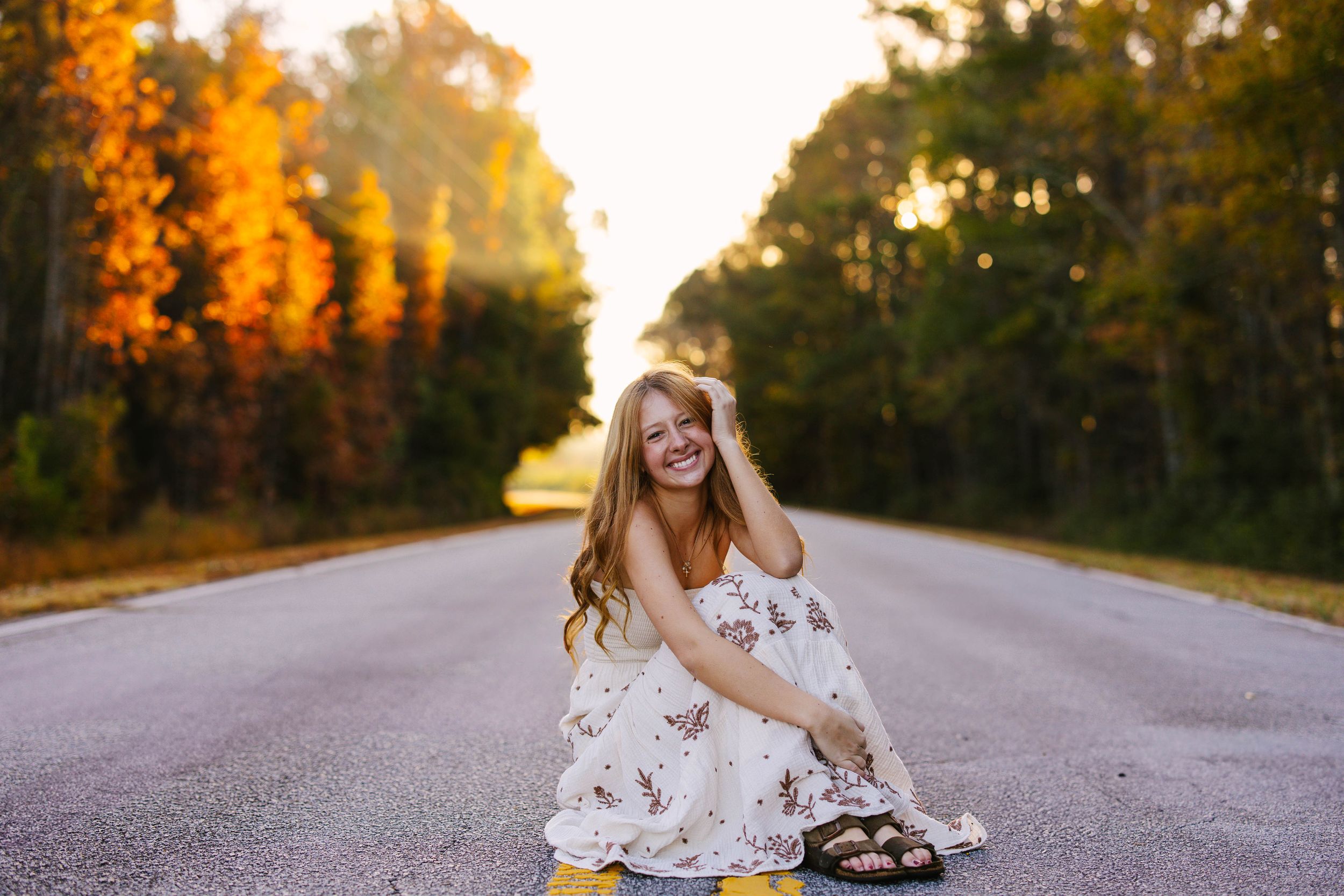 Person in white dress sitting on empty road surrounded by autumn trees during golden hour.