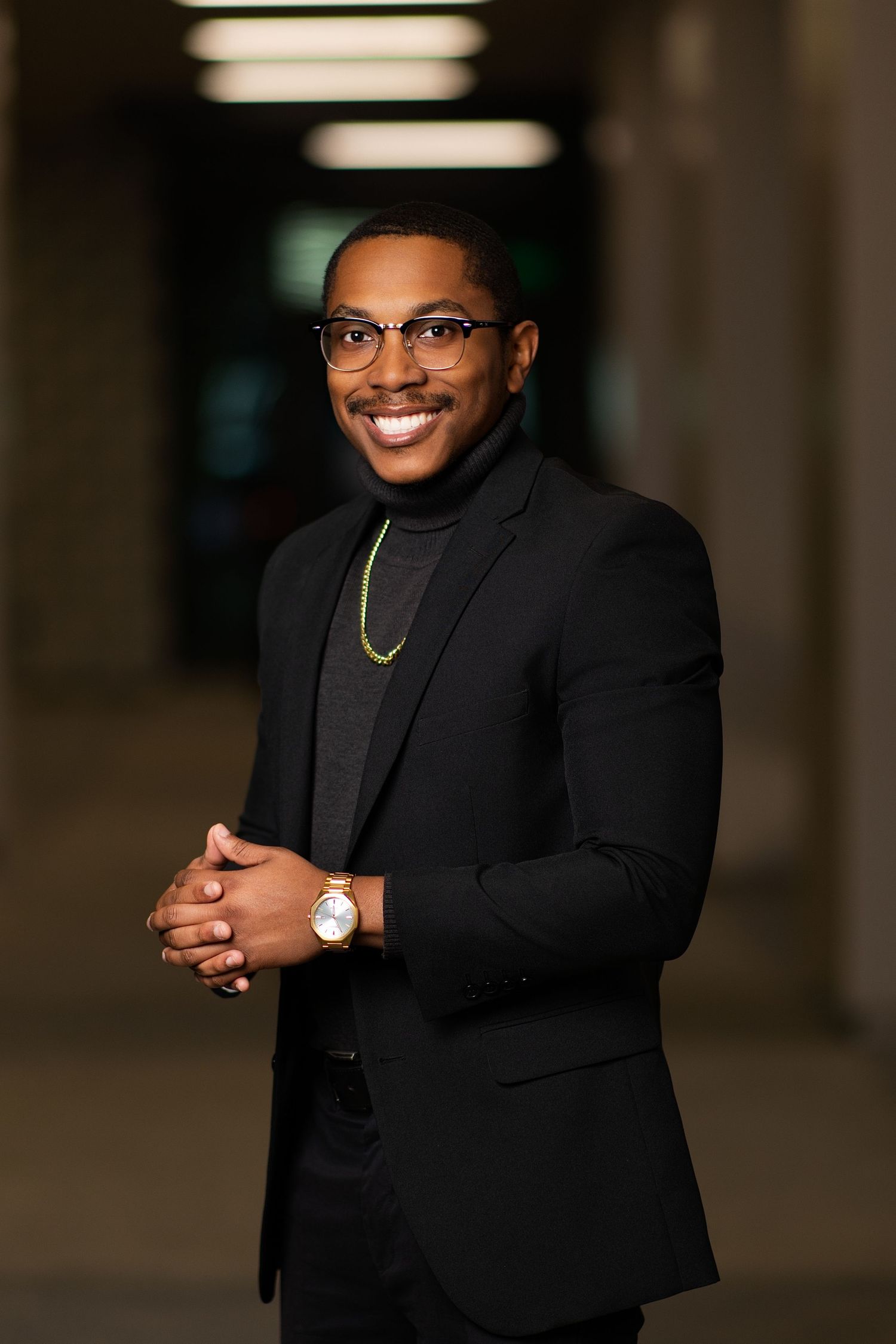 Smiling young Black man in black blazer and turtleneck with glasses and gold watch poses confidently indoors.