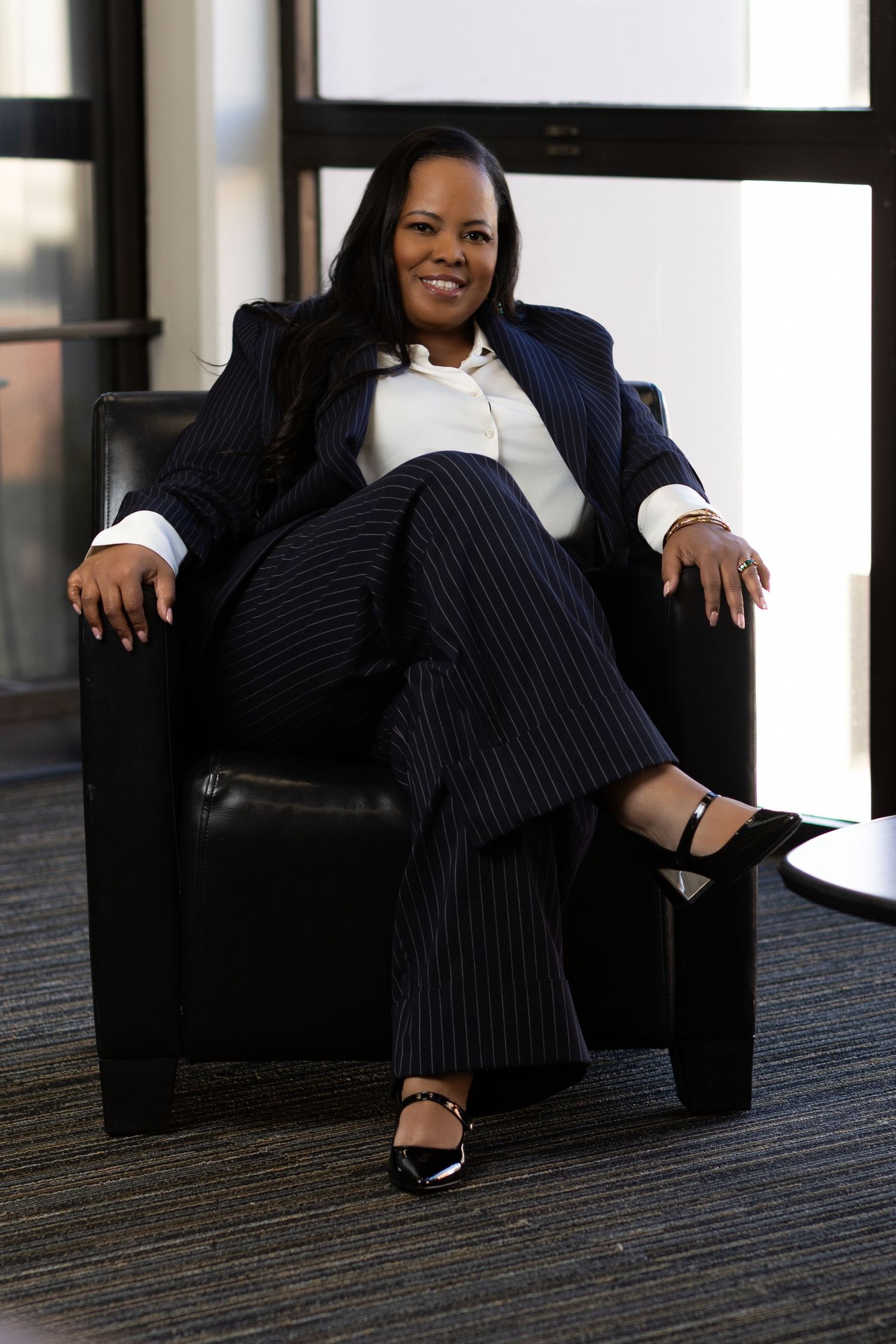Professional woman in pinstripe suit smiling confidently while seated in black leather chair in modern office setting.