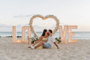 Couple kissing on beach beside illuminated LOVE letters and floral heart arch at sunset.