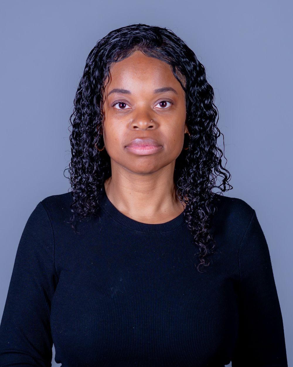 Woman with long curly dark hair wearing a black long-sleeve top poses for a professional headshot against a plain gray background.