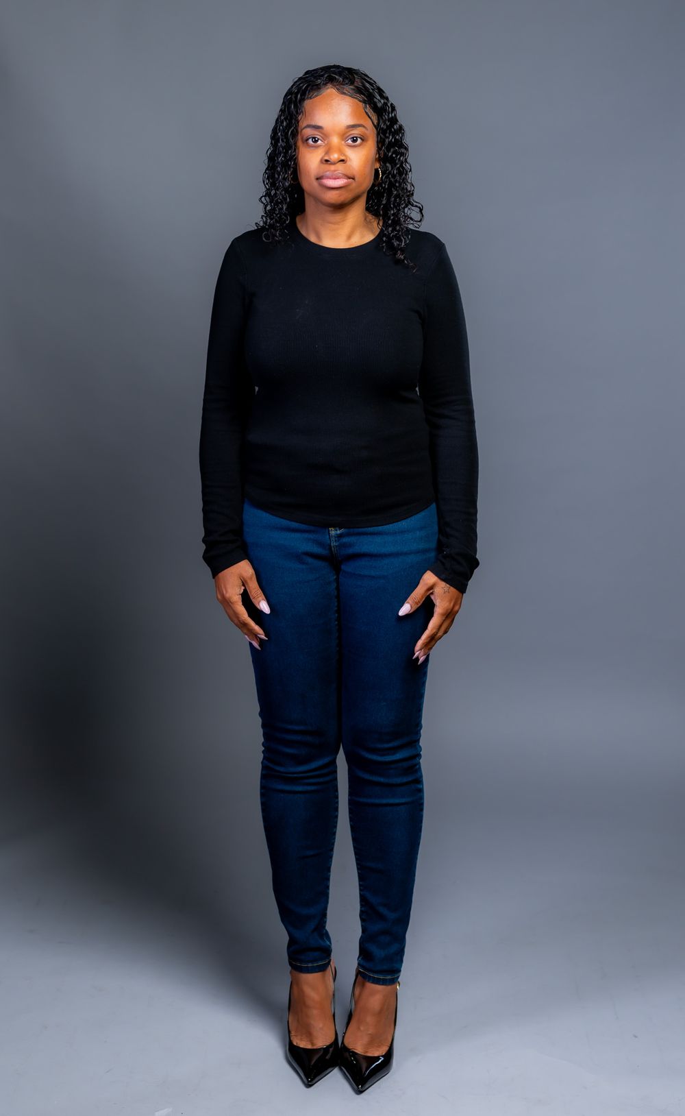 Woman with long curly black hair stands against a gray studio backdrop in a black sweater, dark blue jeans and black pointed-toe heels.