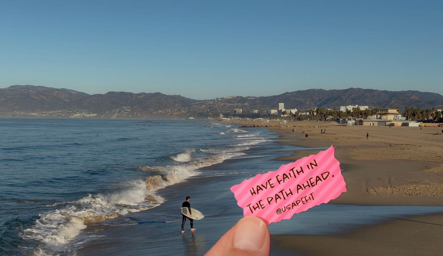 Fingers hold a pink paper note reading Have faith in the path ahead over a sunny beach with a surfer, ocean waves and distant mountains.
