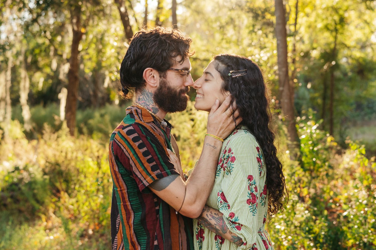 Bearded man in a striped shirt and woman in a floral dress share an affectionate moment, touching faces in a sunlit forest.