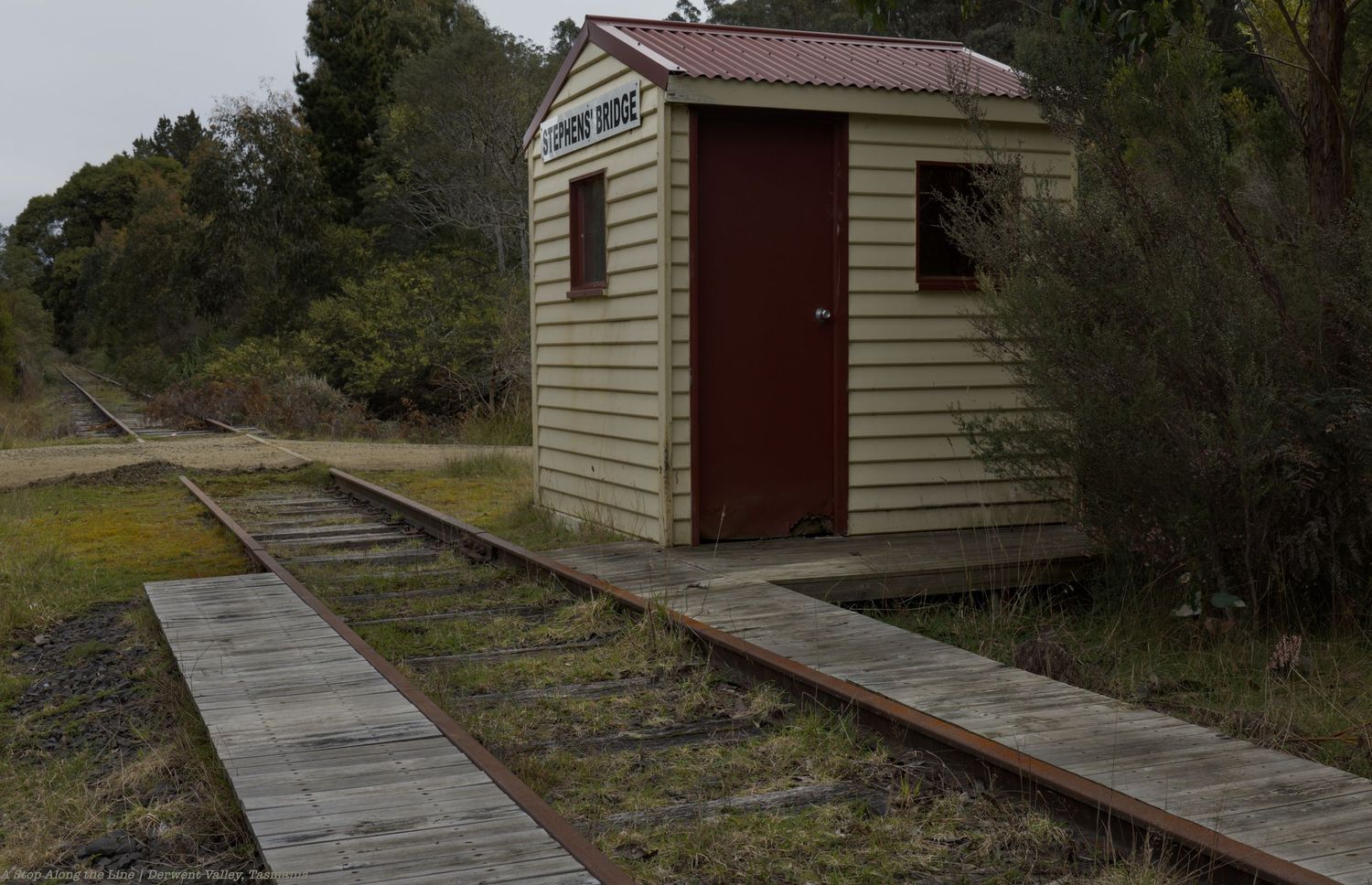 Small weathered wooden railway shelter with a red door beside old train tracks and a timber platform, surrounded by green bushes.
