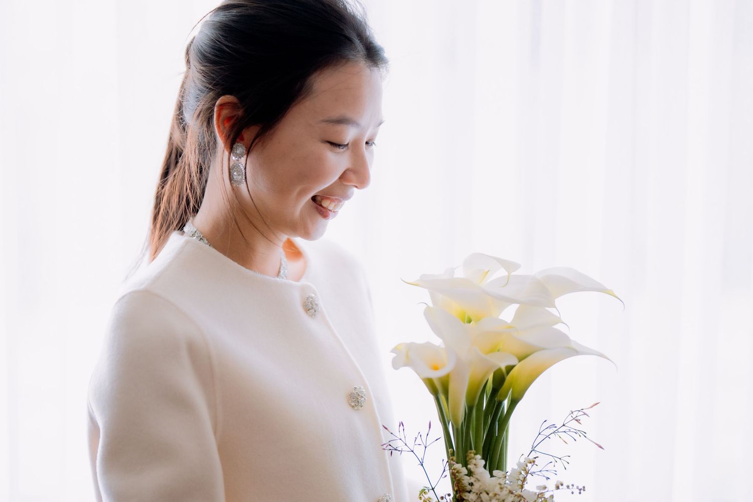 Bride in a cream jacket admires her white calla lily bouquet in window light - intimate elopement photography Sydney