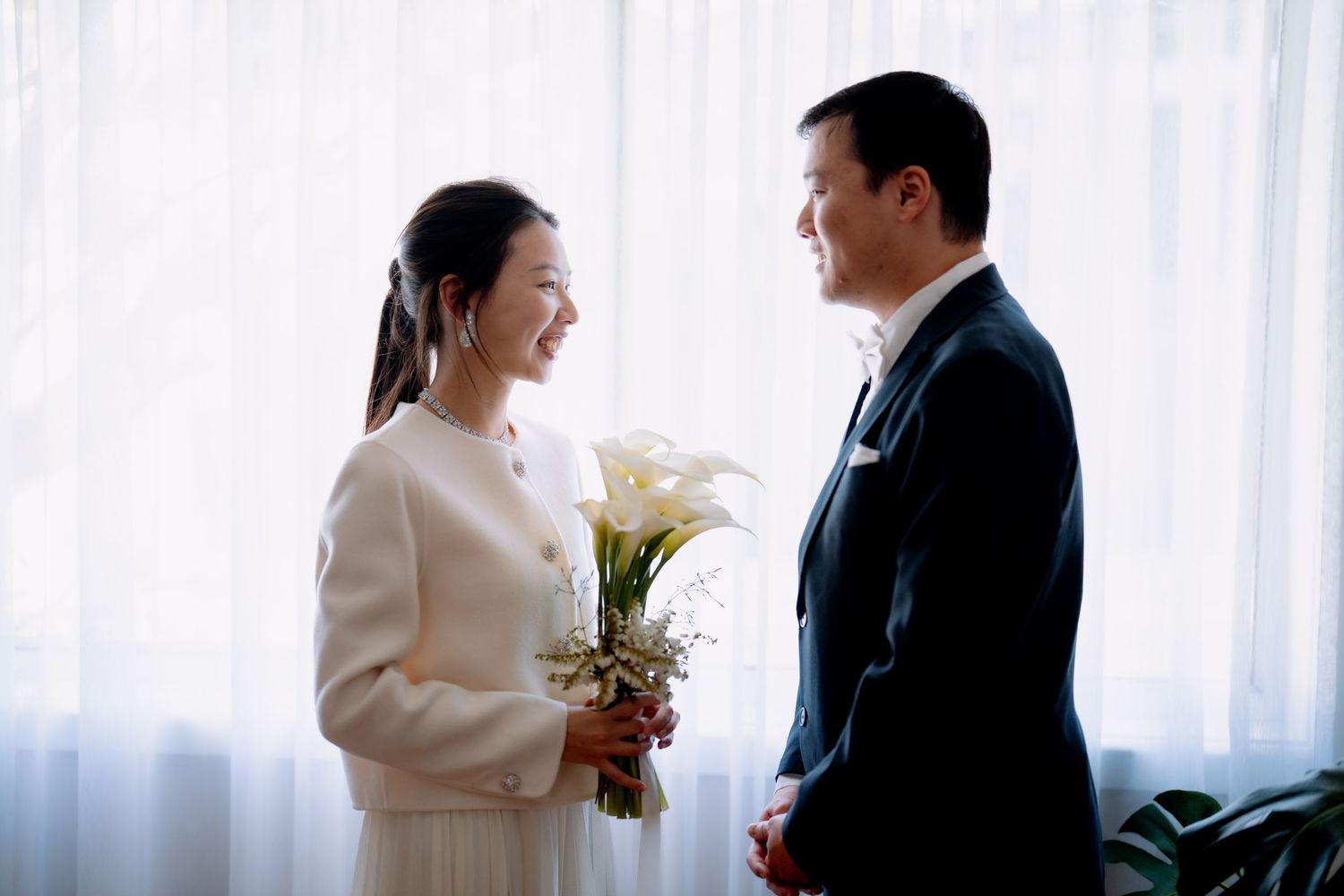 Bride holding calla lilies faces her groom in a dark suit by sheer curtains - small home elopement ceremony