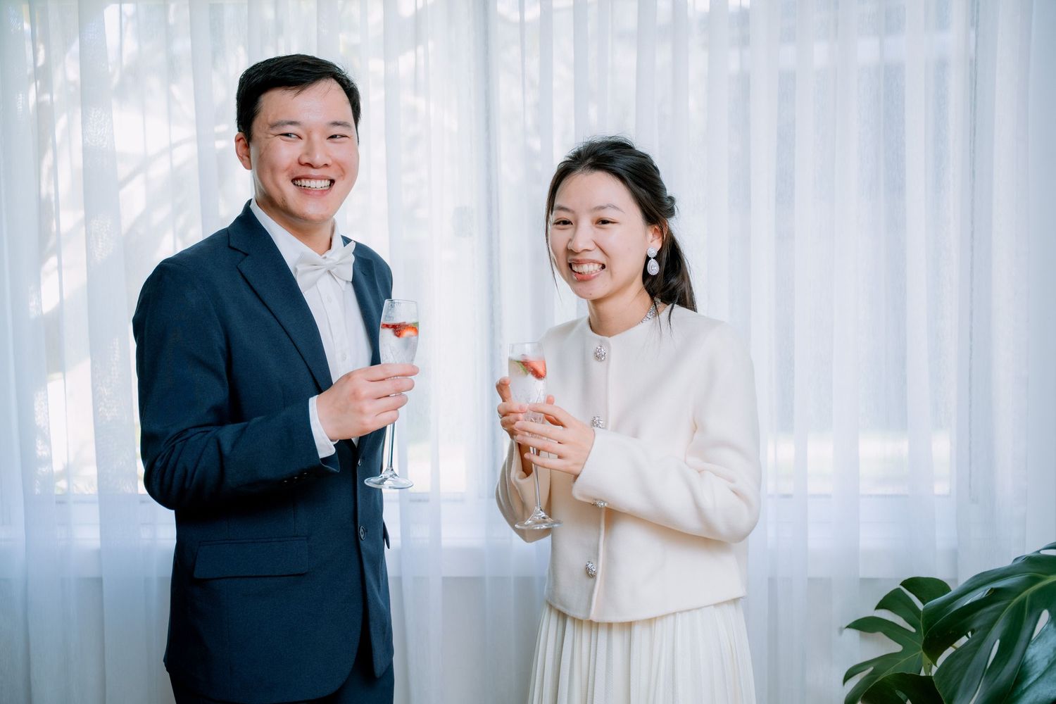 Newlyweds holding champagne glasses by sheer curtains - post-ceremony toast at an intimate elopement