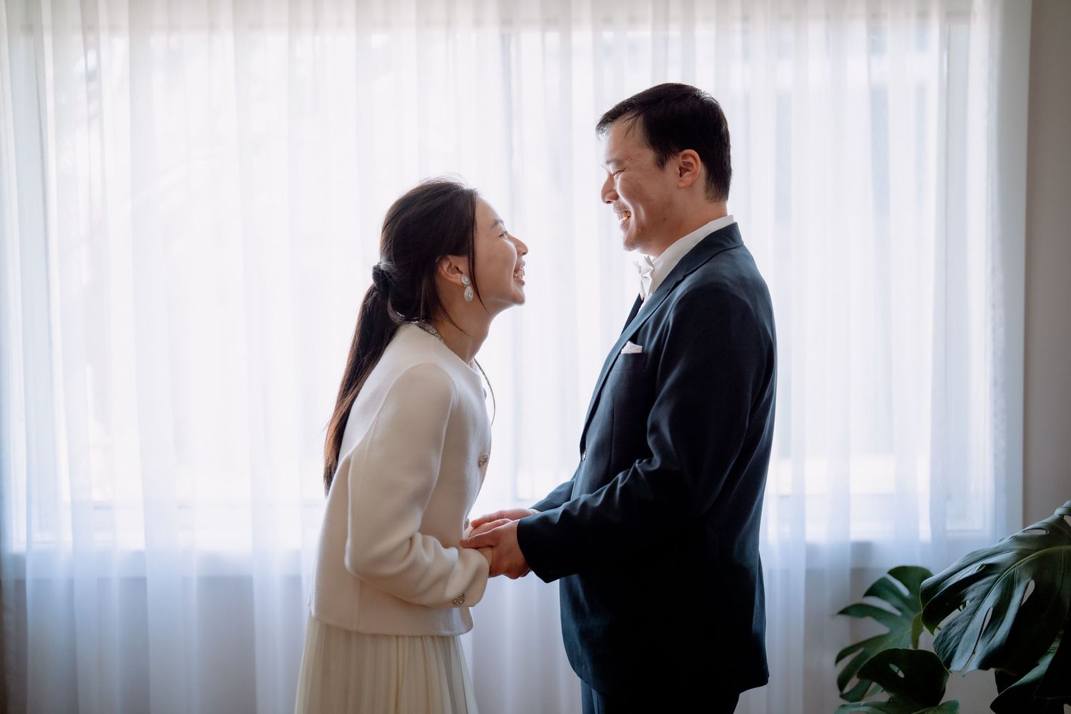 Bride in a cream jacket and groom in a dark suit hold hands by a sunlit window - romantic elopement portrait