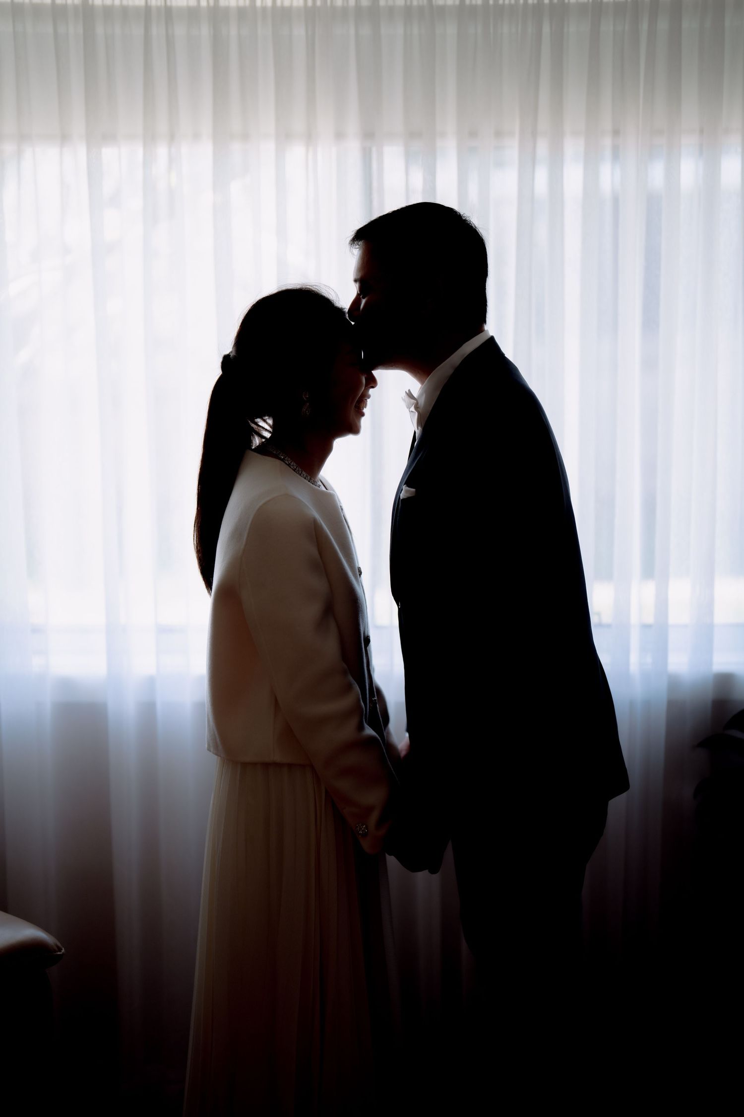 Silhouette of the groom kissing his bride's forehead against a sheer curtain - intimate elopement portrait