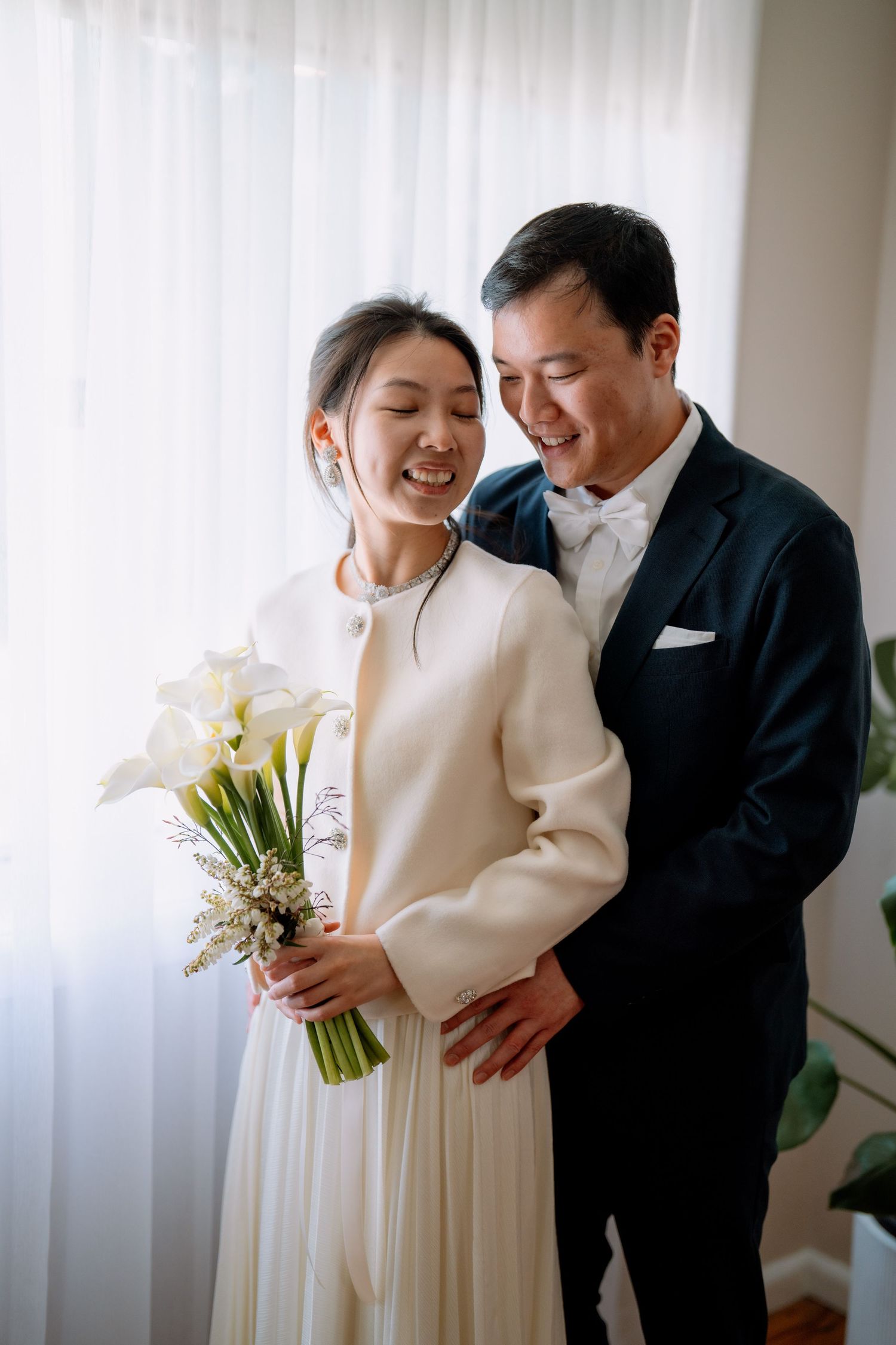 Groom in a navy suit and bow tie hugs his bride from behind as she holds calla lilies - Sydney elopement couple portrait