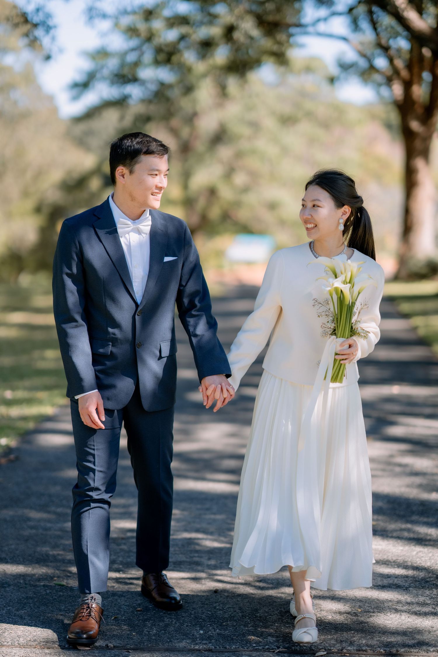 Bride with calla lilies walks with her groom in a navy suit on a sunny path - elopement portraits in the park