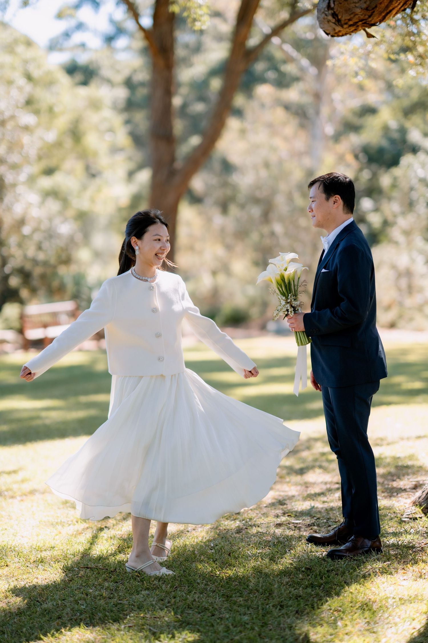 Bride twirls on sunlit grass while her groom holds the calla lily bouquet - fun, candid elopement photos