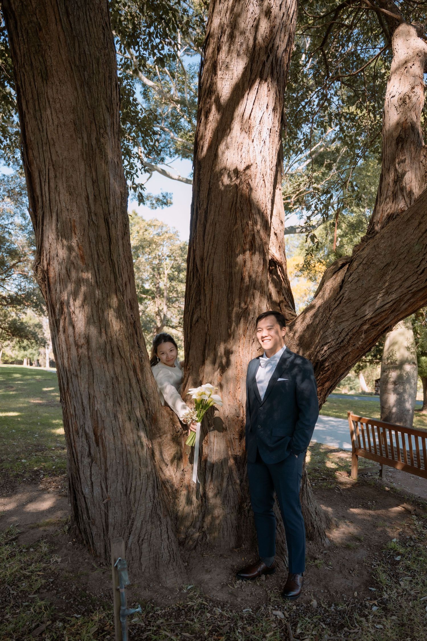 Bride peeks out from behind a tree trunk beside her smiling groom - playful elopement photography